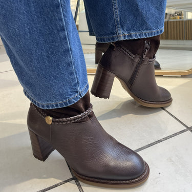 Dark brown leather ankle boots with block heels and braided trim, shown on a person wearing blue jeans in a store setting.