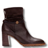 A dark brown ankle boot with a textured pebbled leather lower section, a suede-like folded cuff, a braided strap detail with a gold-tone circular emblem, and a chunky stacked block heel, shown against a plain white background.