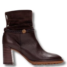 A dark brown ankle boot with a textured pebbled leather lower section, a suede-like folded cuff, a braided strap detail with a gold-tone circular emblem, and a chunky stacked block heel, shown against a plain white background.