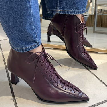 A person wearing blue jeans and burgundy lace-up pointed-toe ankle boots with block heels, standing on a light tiled floor in a store.