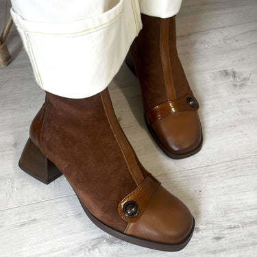Brown ankle boots with a block heel worn with white pants on a light wooden floor.