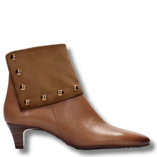 A brown leather ankle boot with a pointed toe, a stacked block heel, and a wide suede-like flap on the side decorated with metallic gold studs.