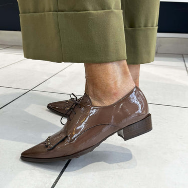 Brown patent leather shoes worn with green pants on a light tiled floor.