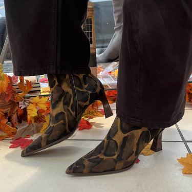 A person wearing pointed-toe ankle boots with a brown and black leopard print pattern, paired with dark brown pants, standing on a light tiled floor with autumn leaves scattered around.