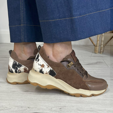 Brown and beige sneakers with cow print details worn by a person in blue pants.