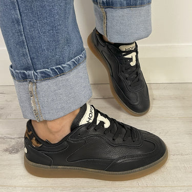 Black sneakers with brown soles worn with rolled-up jeans on a light wooden floor.