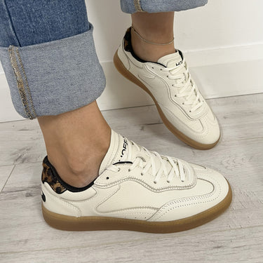 White sneakers with brown soles worn by a person on a wooden floor.