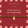 A gold beaded bracelet with a small acorn charm, presented on a pink and red Joma Jewellery gift card that reads 'fresh starts & new beginnings'.