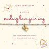 A Joma Jewellery bracelet on a cream-colored display card. The bracelet features a white beaded line with gold and pink details and a gold envelope charm with a red heart.