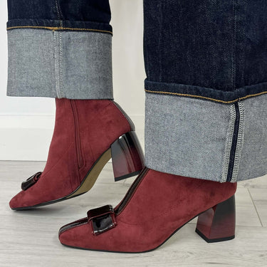 A pair of burgundy suede ankle boots with dark block heels and square buckle details, shown worn with dark cuffed jeans on a light wood floor.