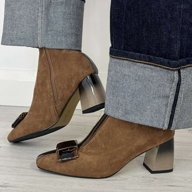 A person wearing brown suede ankle boots with square toes, low block heels, and a small front buckle detail, paired with dark cuffed jeans on a light wood floor.