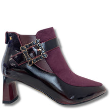 A women's ankle boot with a glossy dark burgundy patent leather toe and heel, a deep burgundy suede upper, and a wide black strap across the instep featuring a decorative gold-tone buckle.