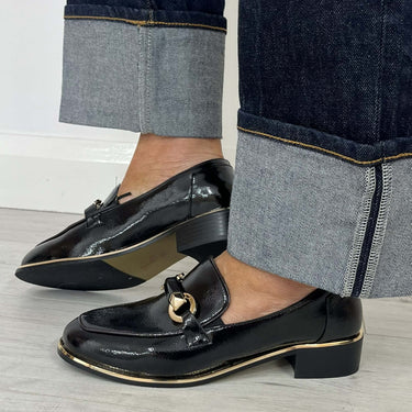 A person wearing black patent leather loafer-style shoes with a gold-tone horsebit detail and low block heels, paired with dark cuffed jeans on a light wood floor.