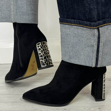 Black suede ankle boots with block heels and leopard-print side embellishments, shown worn with dark cuffed jeans on a light wood floor.