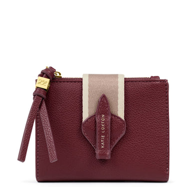 A burgundy pebbled leather zip-around wallet by Katie Loxton, featuring a vertical beige and white stripe on the front, a matching leather tab with the brand name, and a gold-tone zipper pull.
