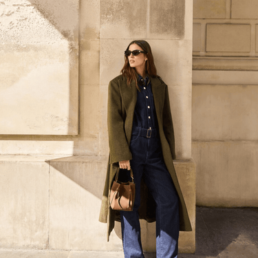 Woman in a green coat and blue outfit standing against a beige wall.
