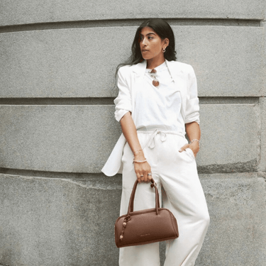 Woman in a white outfit holding a brown handbag against a gray stone wall.