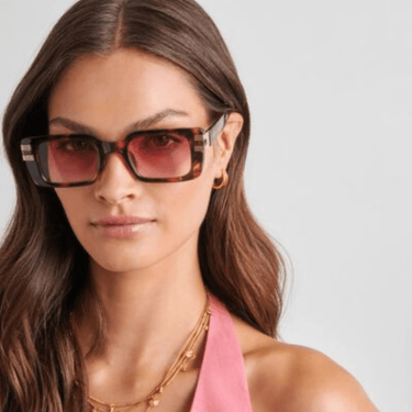 Woman wearing tortoiseshell sunglasses with a pink top against a gray background