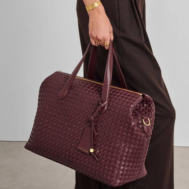 A person in a dark brown dress holds a woven burgundy leather tote bag with gold-tone hardware and a hanging tassel tag against a light gray studio background.