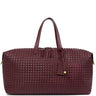 A woven leather duffel bag in a deep burgundy color, featuring dual top handles, a zippered main compartment, and a hanging leather tag with a gold accent, set against a plain white background.