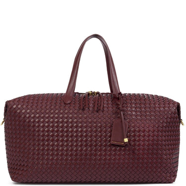 A woven leather duffel bag in a deep burgundy color, featuring dual top handles, a zippered main compartment, and a hanging leather tag with a gold accent, set against a plain white background.