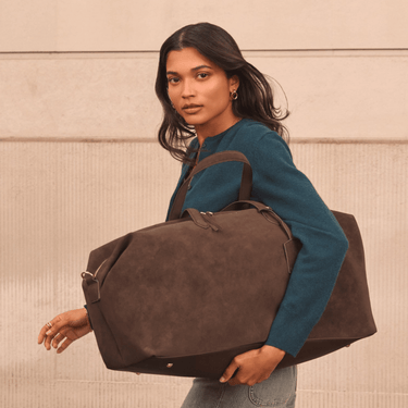 Woman holding a brown suede duffel bag against a beige wall.