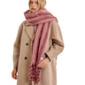 A woman wearing a beige double-breasted coat and a thick, chunky knit scarf with horizontal stripes in dusty rose and burgundy, set against a plain white background.