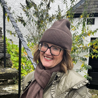 Woman in a green puffer coat and brown hat standing outdoors with stone steps and foliage in the background.