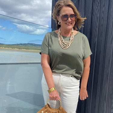 Woman in green shirt and white pants holding a brown bag with gold studs, standing by a glass wall with a scenic view.