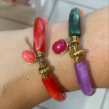 A close-up of a person's wrist wearing two colorful stone bracelets with gold-toned spacers. One bracelet is translucent red with a pink stone accent, and the other is translucent green with a purple stone accent.