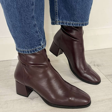 A pair of dark brown leather ankle boots with block heels, shown on a model wearing blue jeans against a light-colored wall and wood-look floor.