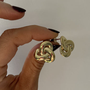 A close-up of a hand with dark nail polish holding a pair of gold-toned interlocking knot stud earrings against a plain light background.