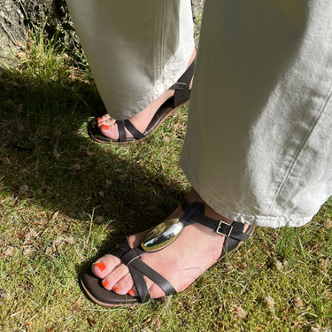 Person wearing sandals and light-colored pants standing on grass next to a tree.