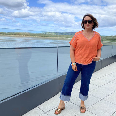 Woman in an orange top and blue jeans standing on a balcony with a scenic view.