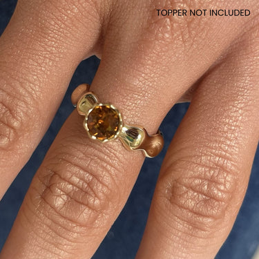 A close-up of a person's hand wearing a gold ring with a round amber-orange center stone. The ring has sculptural, organic-shaped side settings.