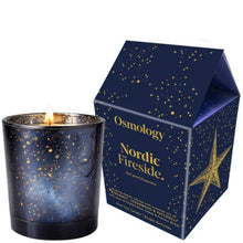 A lit Osmology Nordic Fireside candle in a dark blue speckled glass holder, shown next to its matching house-shaped packaging box with gold star and text details on a white background.