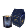 A lit Osmology candle in a dark blue glass vessel with gold speckles, shown next to its matching house-shaped packaging box featuring a gold star and gold lettering on a white background.