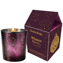 A lit Osmology Winter Berry scented candle in a dark purple glass holder with gold and green speckles, shown next to its matching house-shaped packaging box.