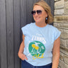 Woman wearing a light blue t-shirt with tennis-themed design against a wooden and stone wall.