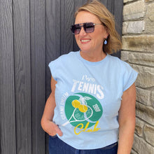 Woman wearing a light blue t-shirt with tennis-themed design against a wooden and stone wall.