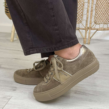 A person wearing dark charcoal pants and taupe-brown suede low-top sneakers with matching laces, standing on a light gray wood-look floor in front of a woven rattan screen.