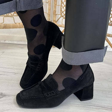 Black suede block-heel loafers worn with sheer black polka-dot tights and dark gray cuffed trousers, shown on a person standing on a light wood floor.