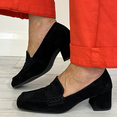 Black suede block-heel loafers worn with bright orange-red cropped pants on a light wood floor.