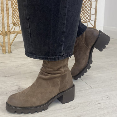 A person wearing dark brown suede ankle boots with chunky lug soles and dark jeans, standing on a light wood floor in front of a gold wicker screen.