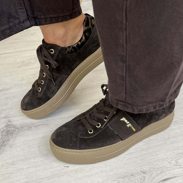 A person wearing dark brown suede low-top sneakers with beige soles and gold-tone eyelets, paired with dark charcoal pants, standing on a light gray wood floor.
