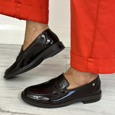 A person wearing bright red trousers and glossy dark brown leather penny loafers, standing on a light gray wood floor against a white wall.