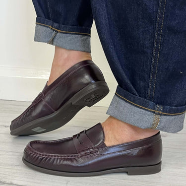 A person wearing dark brown leather penny loafers with cuffed dark blue jeans, standing on a light gray wood floor against a white wall.