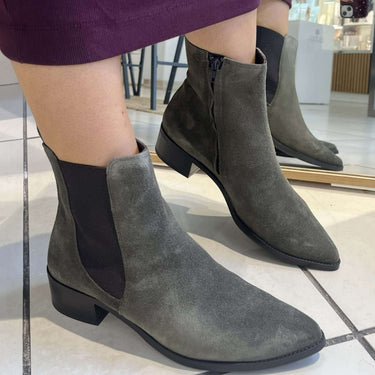 A person wearing gray suede ankle boots with black side panels and low block heels, standing on a light tiled floor in a store.