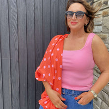Woman wearing a pink top and orange polka dot cardigan against a stone wall.