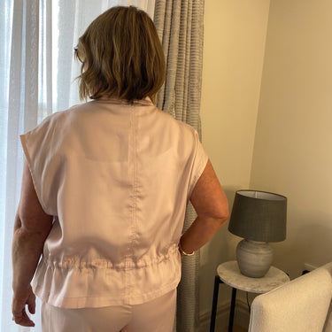 Person wearing a beige sleeveless top and pants standing in a room with a lamp and curtain.